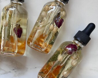 May include: Three clear glass bottles filled with a golden liquid and botanical elements, including dried flowers and orange slices. Each bottle has a black dropper cap. The bottles are arranged on a white marble surface.
