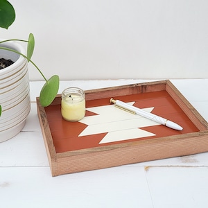 May include: A wooden tray with a geometric design painted in white and orange. A small candle and a pen are on the tray.