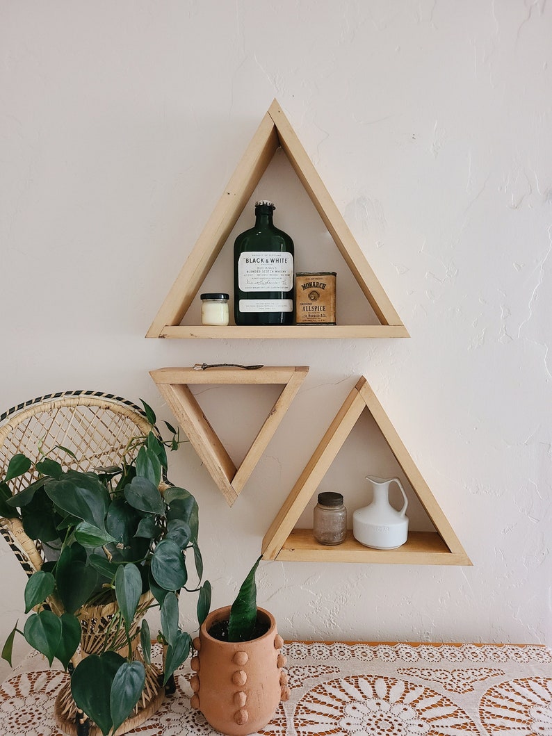 May include: Three triangular wooden shelves mounted on a white wall. The top shelf has a green glass bottle labeled "Black & White Blended Scotch Whisky", a small white candle, and a small tin labeled "Monarch Allspice". The middle shelf is empty. The bottom shelf has a small glass jar and a white ceramic pitcher.