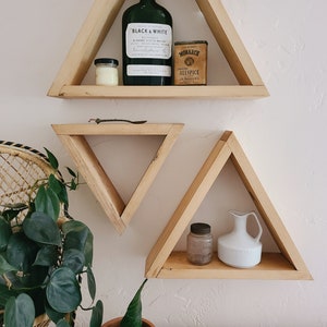 May include: Three triangular wooden shelves mounted on a white wall. The top shelf has a glass bottle with a label that reads "Black & White" and a small tin that reads "Monarch Allspice". The middle shelf is empty. The bottom shelf has a small glass jar and a white ceramic pitcher.