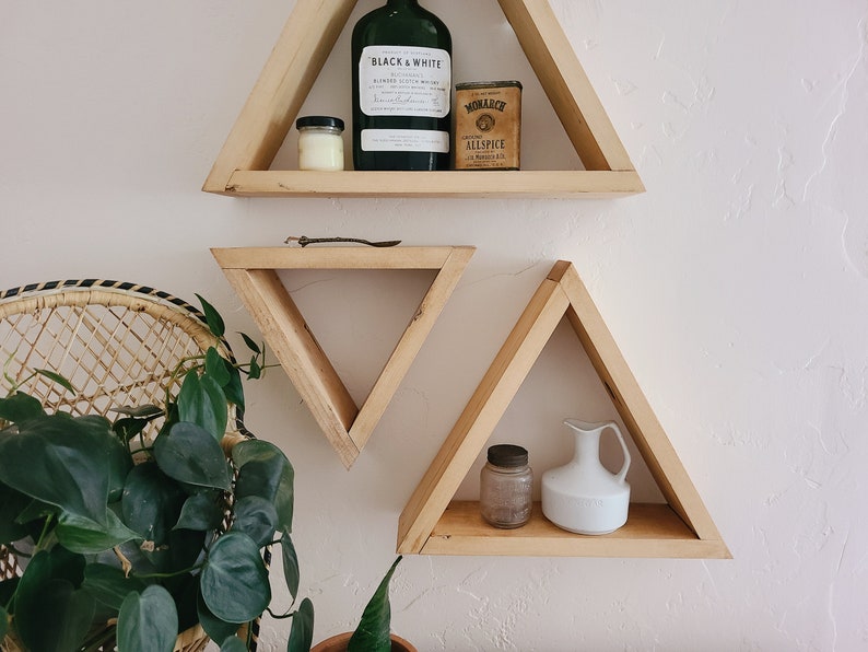 May include: Three triangular wooden shelves mounted on a white wall. The top shelf has a glass bottle with a label that reads "Black & White" and a small tin that reads "Monarch Allspice". The middle shelf is empty. The bottom shelf has a small glass jar and a white ceramic pitcher.