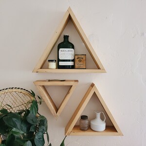 May include: Three wooden triangle shelves mounted on a white wall. The top shelf has a green glass bottle with a label that says "Black & White", a small white candle, and a small tin can with the text "Monarch Hillspice". The middle shelf is smaller and empty. The bottom shelf has a small glass jar and a white ceramic pitcher.
