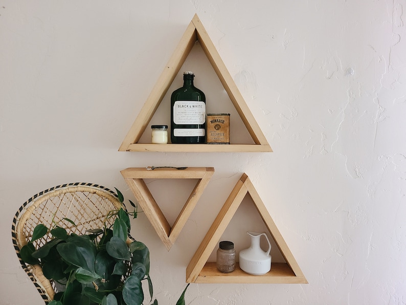 May include: Three wooden triangle shelves mounted on a white wall. The top shelf has a green glass bottle with a label that says "Black & White", a small white candle, and a small tin can with the text "Monarch Hillspice". The middle shelf is smaller and empty. The bottom shelf has a small glass jar and a white ceramic pitcher.