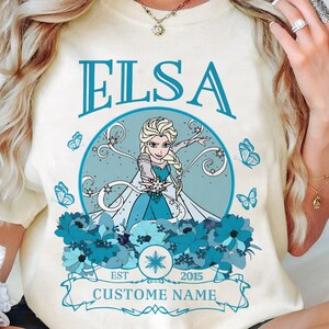 K&ouml;nnte beinhalten: Ein wei&szlig;es T-Shirt mit einer blauen und wei&szlig;en Grafik von Elsa aus Frozen. Die Grafik enth&auml;lt den Text "Elsa", "Est. 2015" und "Custome Name".
