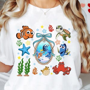 Könnte beinhalten: Weißes T-Shirt mit einem farbenfrohen Cartoon-Design mit Figuren aus dem Film Findet Nemo. Das Design beinhaltet Dory, Marlin, Nemo, eine Schildkröte, einen Oktopus und einen Kugelfisch.