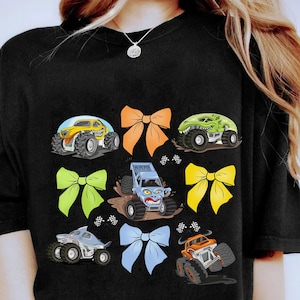 May include: Black t-shirt with a colorful graphic design featuring monster trucks and bows. The trucks are yellow, green, blue, and gray. The bows are orange, green, blue, and yellow.