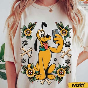Op de afbeelding: Ivoor kleurig T-shirt met een cartoon hond die een selfie maakt. De hond is geel met een rode tong en zwarte oren. Het ontwerp bevat bloemen, botten en pootafdrukken. Het woord "IVORY" staat in een gele rechthoek.