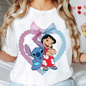 May include: White t-shirt with a blue and purple heart design featuring Stitch and Lilo from Disney's Lilo & Stitch. The heart has a pink and blue bow on top.