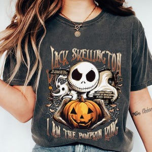 Puede incluir: Camiseta gris oscuro con Jack Skellington sosteniendo una calabaza tallada. La camiseta tiene el texto "Jack Skellington" y "I am the Pumpkin King". El dise&ntilde;o incluye a Zero, telara&ntilde;as y el texto "Christmas time is buzzing in my skull."