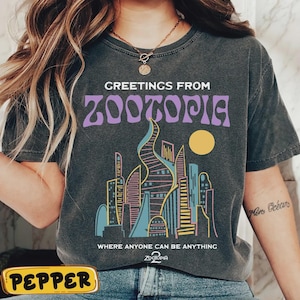 May include: Dark gray t-shirt with the text "GREETINGS FROM ZOOTOPIA" in purple, above a colorful cityscape illustration. The shirt also features the phrase "WHERE ANYONE CAN BE ANYTHING" and the Zootopia logo.
