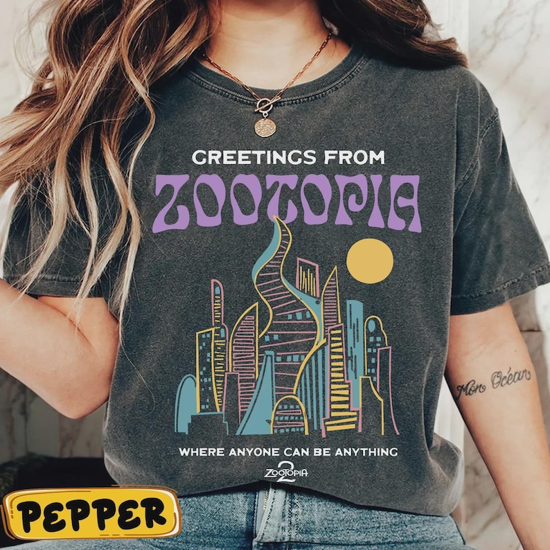 May include: Dark gray t-shirt with the text "GREETINGS FROM ZOOTOPIA" in purple, above a colorful cityscape illustration. The shirt also features the phrase "WHERE ANYONE CAN BE ANYTHING" and the Zootopia logo.