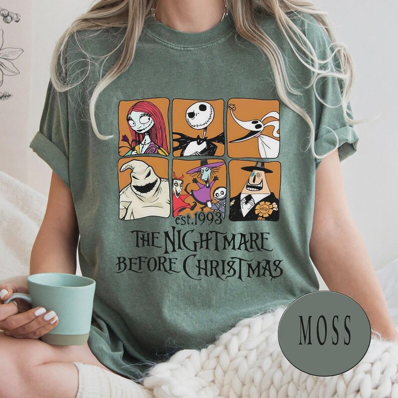 May include: A moss green t-shirt featuring a graphic design from "The Nightmare Before Christmas." The design includes six character portraits in orange squares and the text "est. 1993" and "THE NIGHTMARE BEFORE CHRISTMAS" in black. The shirt has rolled-up sleeves.