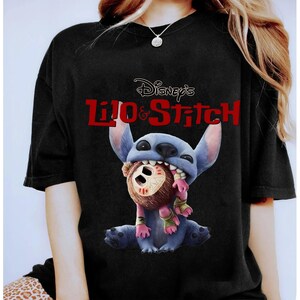 Może przedstawiać: Czarny t-shirt z grafiką Stitcha z filmu Disneya Lilo & Stitch. Niebieski kosmita jest przedstawiony z brązową lalką w ustach. Napis "Disney's Lilo & Stitch" jest wydrukowany na czerwono nad postacią.