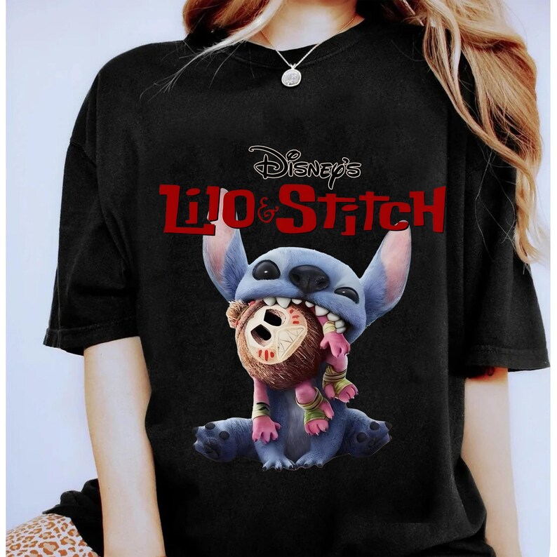 Może przedstawiać: Czarny t-shirt z grafiką Stitcha z filmu Disneya Lilo & Stitch. Niebieski kosmita jest przedstawiony z brązową lalką w ustach. Napis "Disney's Lilo & Stitch" jest wydrukowany na czerwono nad postacią.
