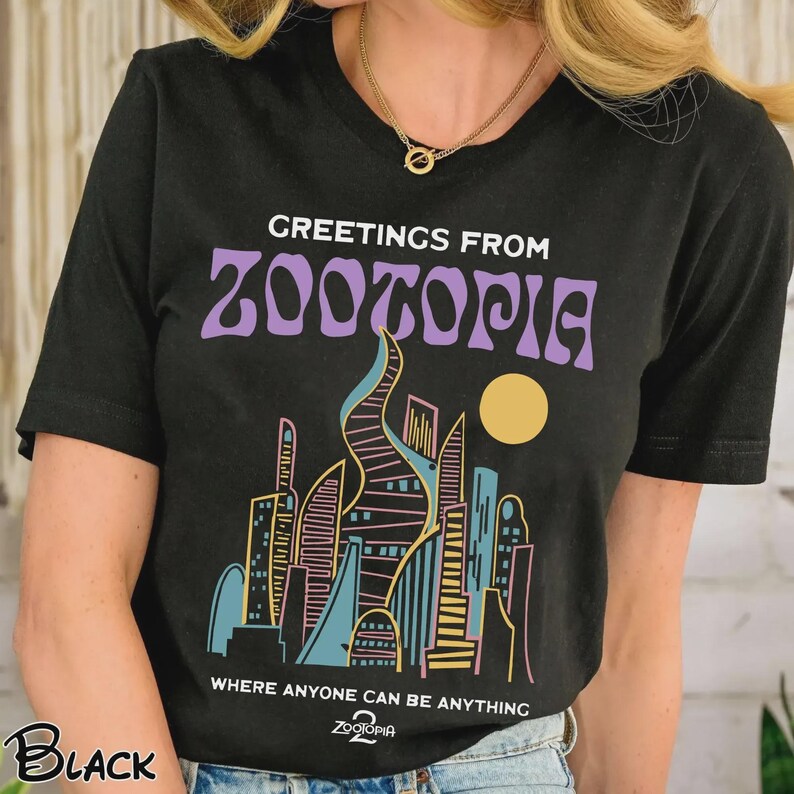 May include: Black t-shirt with a colorful graphic of a cityscape and the text "GREETINGS FROM ZOOTOPIA" in purple. Below the graphic, the text reads "WHERE ANYONE CAN BE ANYTHING".