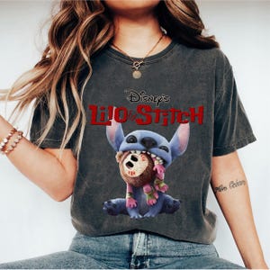 Może przedstawiać: Ciemnoszara koszulka z Lilo & Stitch Disneya. Grafika przedstawia Stitcha, niebieskiego kosmitę, trzymającego brązowo-r&oacute;żową lalkę. Logo Disneya jest czerwone. Koszulka ma swobodny, wygodny styl.
