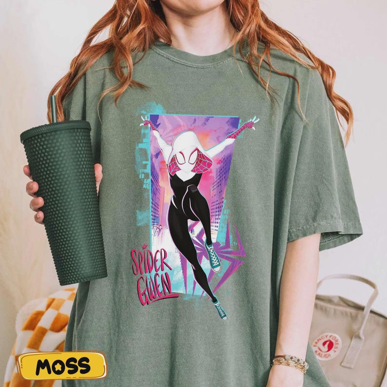 May include: Olive green t-shirt featuring a graphic of Spider-Gwen in a dynamic pose, set against a cityscape backdrop. The design includes the text "Spider Gwen" in a stylized font. The shirt is a relaxed fit.