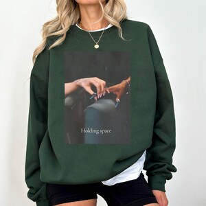 Pu&ograve; includere: Felpa verde scuro con una maglietta bianca che sbuca dallo scollo. La parte anteriore presenta un'immagine rettangolare con le parole "Holding space" e una foto di mani. La felpa ha maniche lunghe e una vestibilit&agrave; ampia.