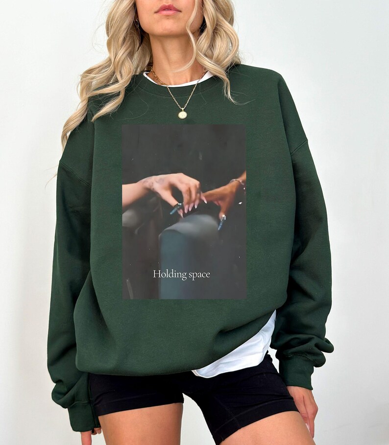 Pu&ograve; includere: Felpa verde scuro con una maglietta bianca che sbuca dallo scollo. La parte anteriore presenta un'immagine rettangolare con le parole "Holding space" e una foto di mani. La felpa ha maniche lunghe e una vestibilit&agrave; ampia.
