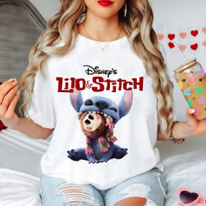 Może przedstawiać: Biała koszulka z logo Disney's Lilo & Stitch w kolorze czerwonym i czarnym. Grafika przedstawia Stitcha, niebieskiego kosmitę, trzymającego brązowego misia. Koszulka ma okrągły dekolt.