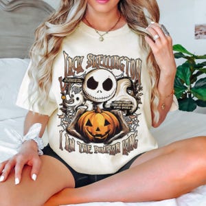 Puede incluir: Camiseta color crema con un dise&ntilde;o de Jack Skellington sosteniendo una calabaza naranja tallada. Se muestra el texto "Jack Skellington" y "I am the Pumpkin King". El dise&ntilde;o incluye telara&ntilde;as y otros elementos tem&aacute;ticos de Halloween.