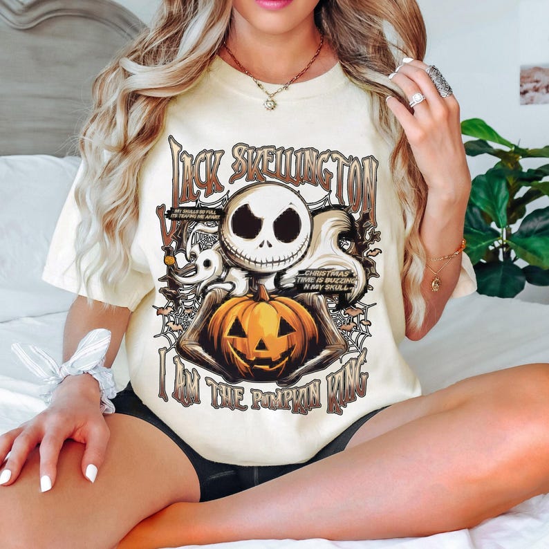 Puede incluir: Camiseta color crema con un dise&ntilde;o de Jack Skellington sosteniendo una calabaza naranja tallada. Se muestra el texto "Jack Skellington" y "I am the Pumpkin King". El dise&ntilde;o incluye telara&ntilde;as y otros elementos tem&aacute;ticos de Halloween.
