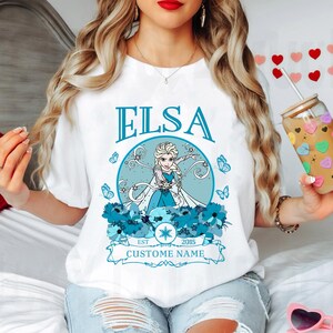 K&ouml;nnte beinhalten: Wei&szlig;es T-Shirt mit einem blauen und wei&szlig;en Grafik von Elsa aus Frozen. Die Grafik enth&auml;lt den Text "Elsa", "Est. 2015" und "Custome Name".