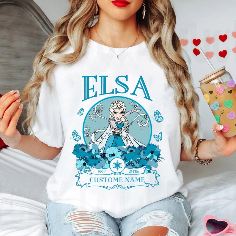 K&ouml;nnte beinhalten: Wei&szlig;es T-Shirt mit einem blauen und wei&szlig;en Grafik von Elsa aus Frozen. Die Grafik enth&auml;lt den Text "Elsa", "Est. 2015" und "Custome Name".