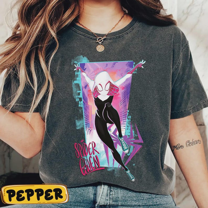 May include: Dark gray t-shirt featuring a graphic of Spider-Gwen in a dynamic pose, set against a cityscape backdrop. The design includes the text "Spider Gwen" in pink and white. The shirt has a vintage, worn look.