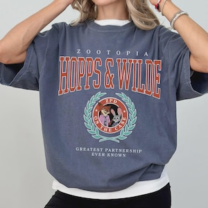 Op de afbeelding: Blauw-grijs Zootopia T-shirt met de tekst "HOPPS & WILDE" in het rood. Het shirt heeft een cirkelvormig embleem met Nick Wilde en Judy Hopps. De tekst "GREATEST PARTNERSHIP EVER KNOWN" staat er ook op.