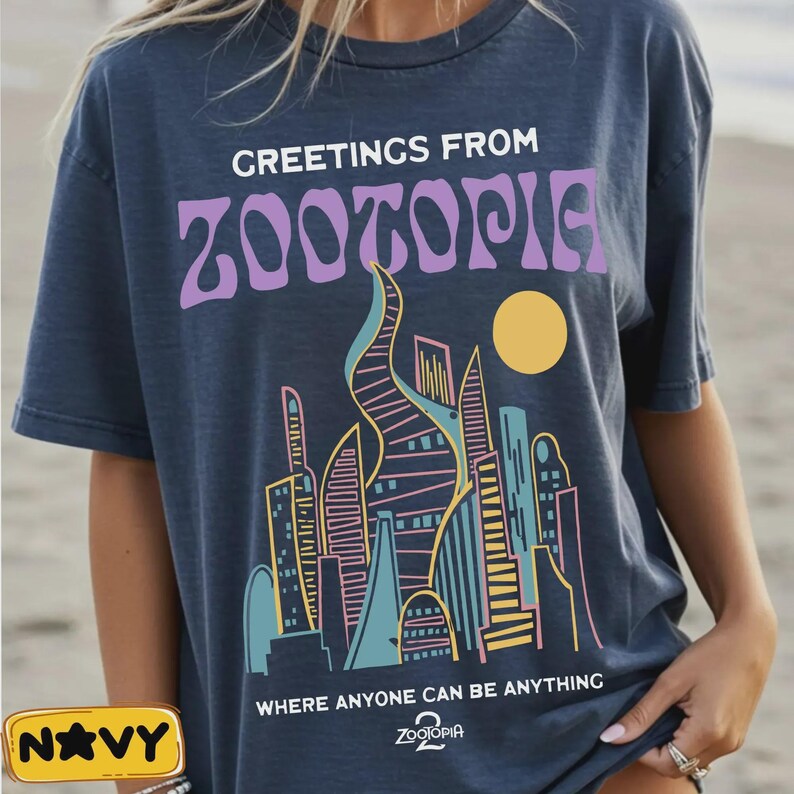 May include: Navy blue t-shirt with the text "GREETINGS FROM ZOOTOPIA" in white and purple. Below the text is a colorful illustration of a city skyline with a yellow sun. The shirt also has the text "WHERE ANYONE CAN BE ANYTHING".