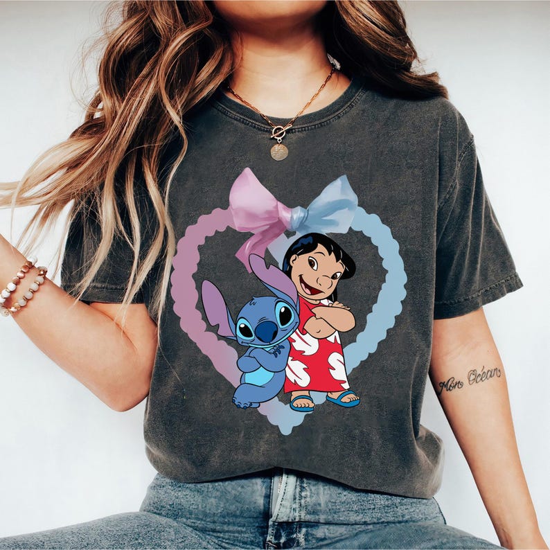 May include: A dark gray t-shirt with a graphic print of Stitch and Lilo from the Disney movie Lilo & Stitch. The graphic features Stitch and Lilo standing together inside a heart-shaped outline with a pink and blue bow.