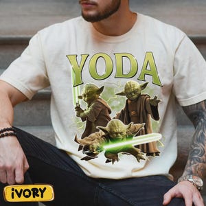 May include: Ivory t-shirt with a graphic of Yoda wielding green lightsabers, and the word "YODA" in green. The design shows three Yoda figures in action poses, with lightning bolts. The t-shirt is ivory in colour.
