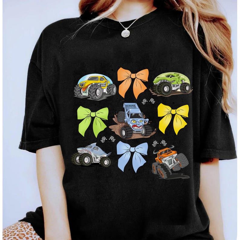 May include: Black t-shirt with a colorful graphic design featuring monster trucks and bows. The trucks are yellow, green, blue, and gray. The bows are orange, green, blue, and yellow.