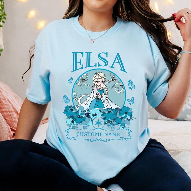 K&ouml;nnte beinhalten: Hellblaues T-Shirt mit einem Aufdruck von Elsa aus Frozen. Der Aufdruck enth&auml;lt einen Schneeflock, Schmetterlinge und den Text "Elsa" und "Custome Name".