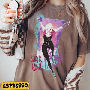 May include: Espresso-colored t-shirt featuring a graphic of Spider-Gwen in a dynamic pose. The design includes a cityscape backdrop and the words "Spider Gwen" in a stylized font. The shirt is a casual wear item.