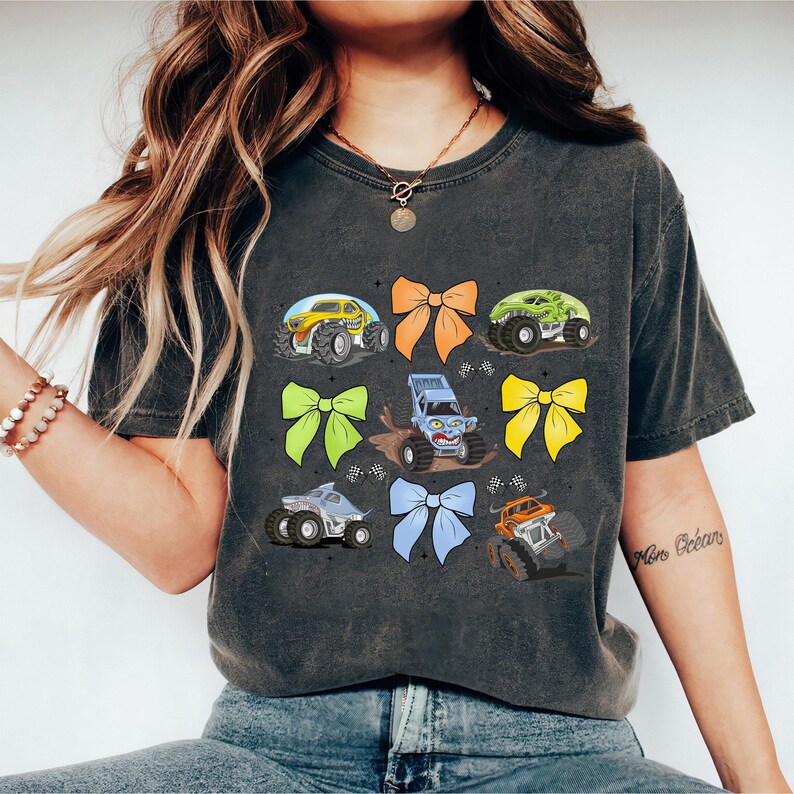 May include: A dark gray t-shirt with a graphic design of monster trucks and colorful bows. The trucks are blue, green, and yellow. The bows are orange, yellow, green, and blue.