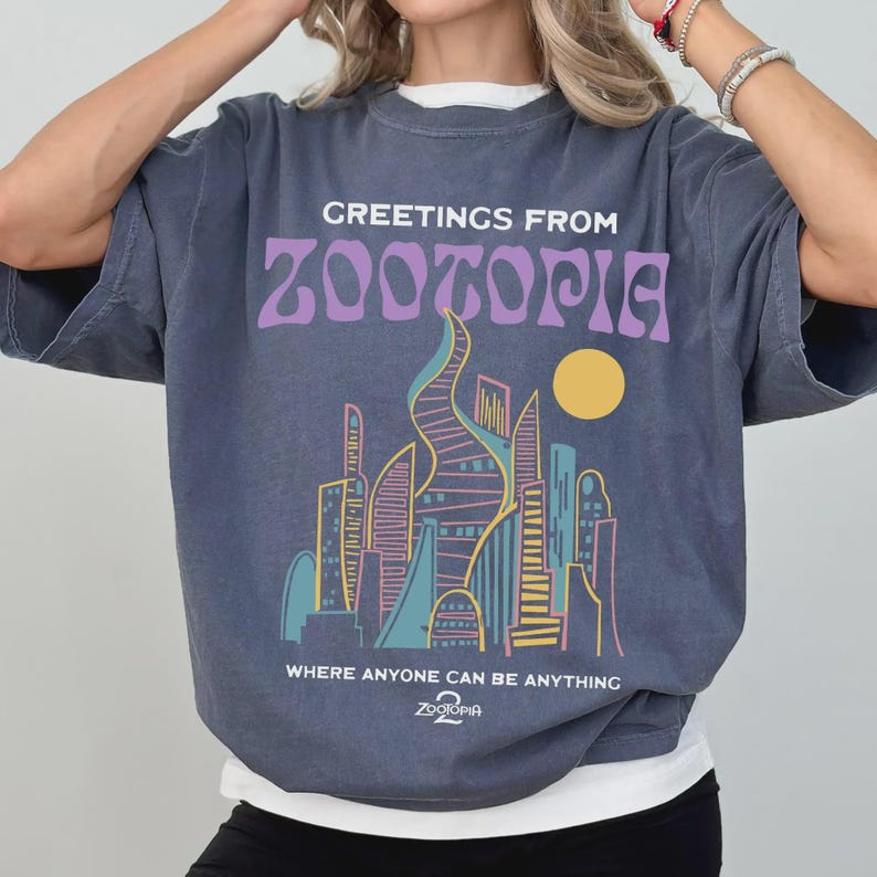 May include: A blue-grey t-shirt with the text "GREETINGS FROM ZOOTOPIA" in purple, above a colorful cityscape illustration. The shirt also features the text "WHERE ANYONE CAN BE ANYTHING" and the Zootopia logo.