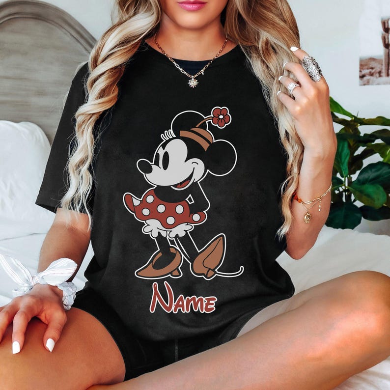 Peut inclure: T-shirt noir avec un motif de Minnie Mouse portant une robe &agrave; pois rouges et blancs. Le texte "Name" est imprim&eacute; sous le motif.