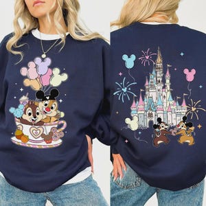 May include: Navy blue sweatshirt featuring Chip and Dale in a teacup with Mickey Mouse balloons. The back of the sweatshirt displays a castle, fireworks, and Chip and Dale. The sweatshirt has a white collar.