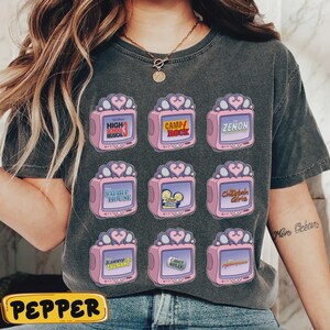 May include: Dark gray t-shirt featuring nine pink television graphics with various titles, including "High School Musical," "Camp Rock," and "Zenon." Each TV has a heart-shaped antenna. The word "PEPPER" is at the bottom.