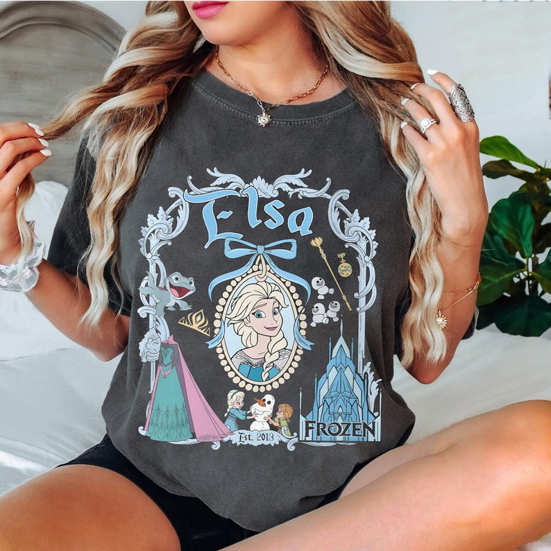 May include: A gray t-shirt with a graphic print of Elsa from Frozen. The graphic includes a portrait of Elsa, a castle, and the text "Elsa" and "Frozen".