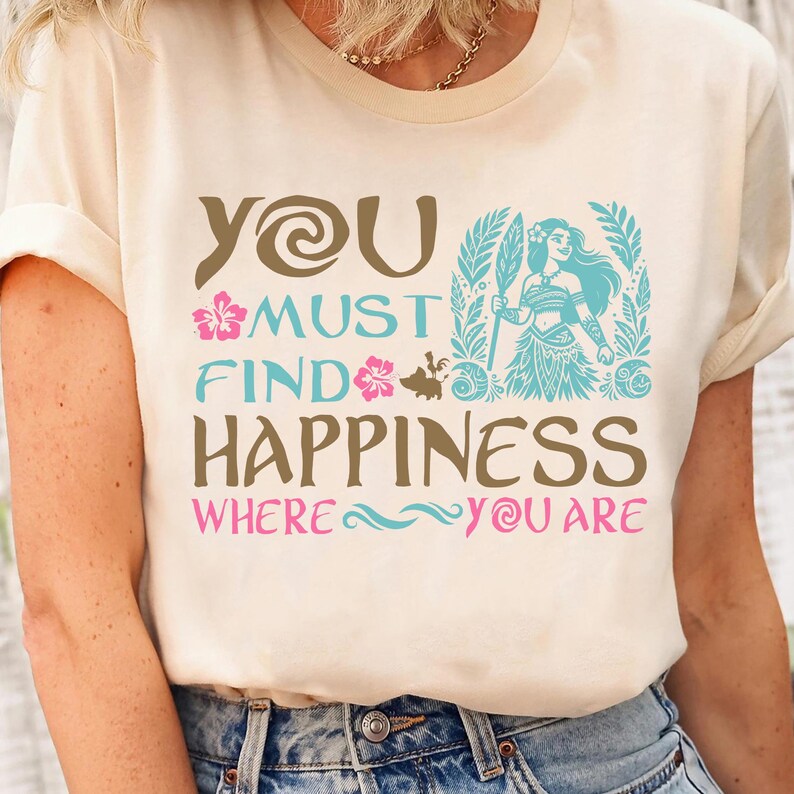 May include: A white t-shirt with a brown graphic that says "You must find happiness where you are". The graphic also includes a cartoon image of a woman with brown hair and a blue dress, a small pig, and pink flowers.
