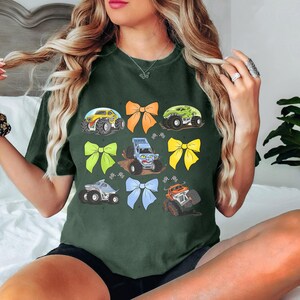 May include: A dark green t-shirt with a graphic print of monster trucks and colorful bows. The trucks are blue, green, and yellow. The bows are yellow, orange, and blue.