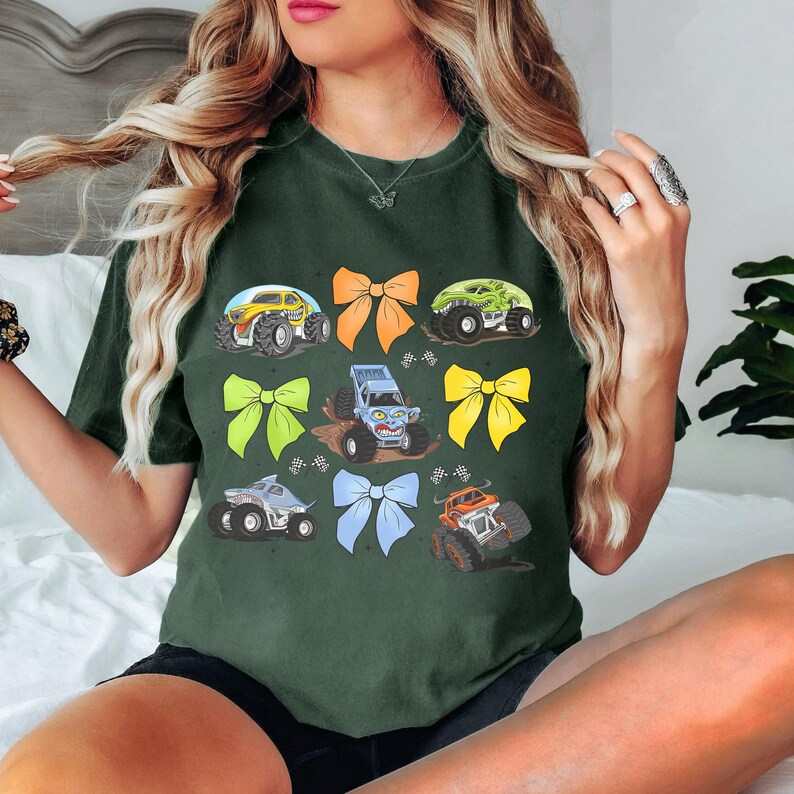 May include: A dark green t-shirt with a graphic print of monster trucks and colorful bows. The trucks are blue, green, and yellow. The bows are yellow, orange, and blue.