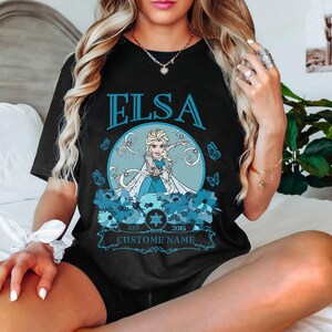 K&ouml;nnte beinhalten: Schwarzes T-Shirt mit einem Aufdruck von Elsa aus Frozen. Der Aufdruck zeigt Elsa mit blauen Haaren und einem blauen Kleid, umgeben von blauen Blumen und Schneeflocken. Der Text "Elsa" befindet sich &uuml;ber dem Aufdruck, und "Est. 2015 Custom Name" befindet sich unter dem Aufdruck.