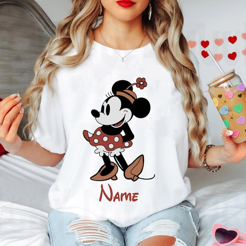 Peut inclure: T-shirt blanc avec une image de dessin anim&eacute; de Minnie Mouse portant une robe &agrave; pois rouges et blancs et des chaussures rouges. Le texte "NAME" est imprim&eacute; sous l'image.
