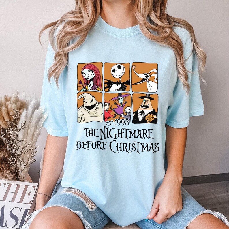 May include: Light blue t-shirt featuring a graphic of characters from "The Nightmare Before Christmas." The design includes six character portraits in orange squares and the text "est.1993 THE NIGHTMARE BEFORE CHRISTMAS" in black.