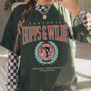 Op de afbeelding: Donkergroen oversized T-shirt met de tekst "ZOOTOPIA HOPPS & WILDE" in rood en wit. Het shirt heeft een cirkelvormig embleem met stripfiguren en de woorden "ON THE CASE". De zin "GREATEST PARTNERSHIP EVER KNOWN" staat eronder.