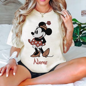 Peut inclure: Un t-shirt blanc avec une image de dessin anim&eacute; de Minnie Mouse de style vintage portant une robe &agrave; pois rouges et des chaussures rouges. Le texte "Name" est imprim&eacute; sous l'image.